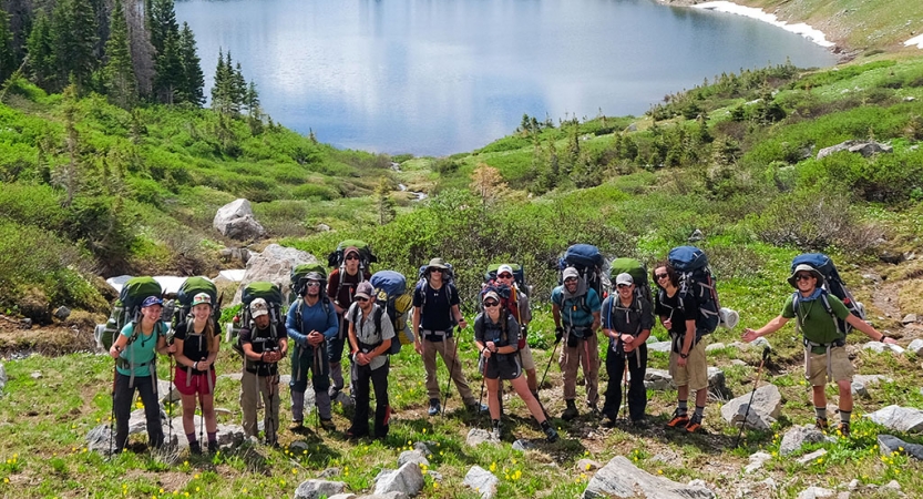 A group of students pose for a photo in front of a small body of water that is surrounded by green grass.